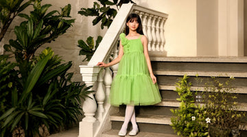 Girl wearing a lime green designer tulle dress with floral shoulder appliqués, standing on elegant stone stairs in a Mediterranean-style courtyard surrounded by lush greenery.
