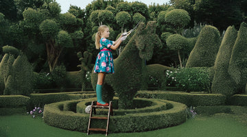 Girl in a vibrant floral dress standing on a ladder, trimming a topiary horse in a beautifully sculpted garden.