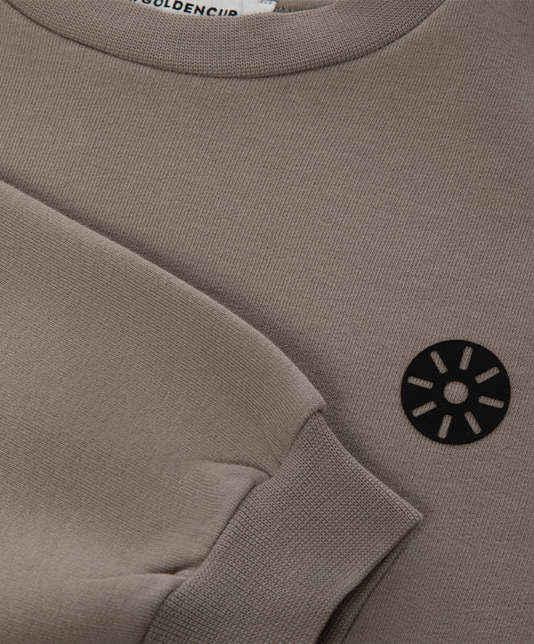 Close-up detail of a taupe girls sweatshirt highlighting the ribbed cuff, crew neckline, soft knit texture, and minimalist chest emblem, designed for everyday wear, school outfits, and casual layering, available in sizes 3–14