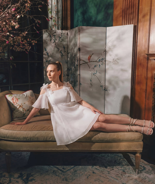 Woman in a white dress sitting on a vintage couch with a floral screen in the background