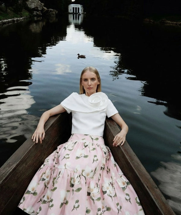 Woman in a white top and pink floral skirt sitting in a wooden boat on a calm lake.