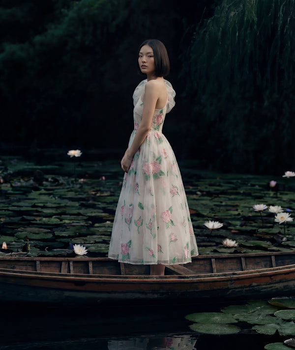 Woman in a floral dress standing in a boat on a pond with lily pads.
