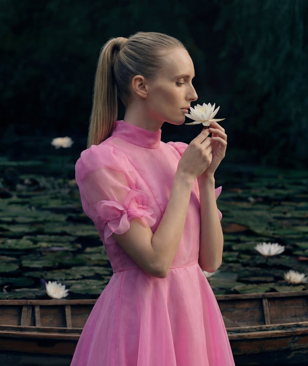 Woman in a pink dress holding a white flower by a body of water with lily pads.