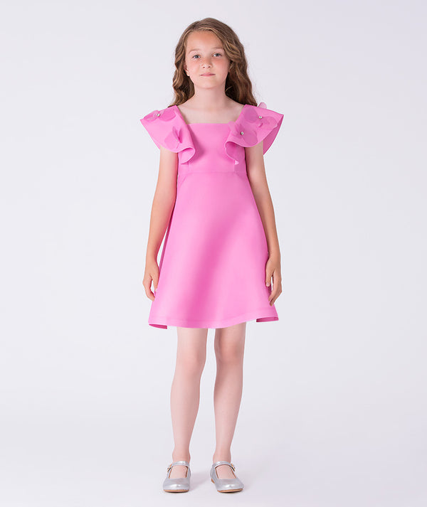 Luxury girls occasion dress in vibrant pink, worn by a 7-year-old girl, featuring sculptural ruffle shoulders, floral appliqués and a softly flared skirt for special events.