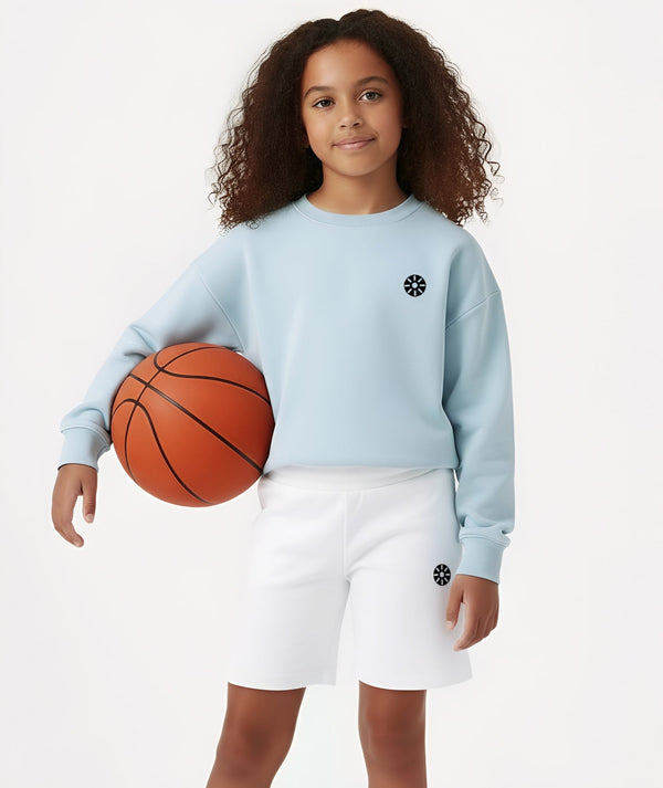 Front-facing lifestyle image of a girl wearing a light blue long sleeve sweatshirt with white shorts while holding a basketball, styled for sporty school days, casual play, and comfortable everyday wear, available in sizes 3–14