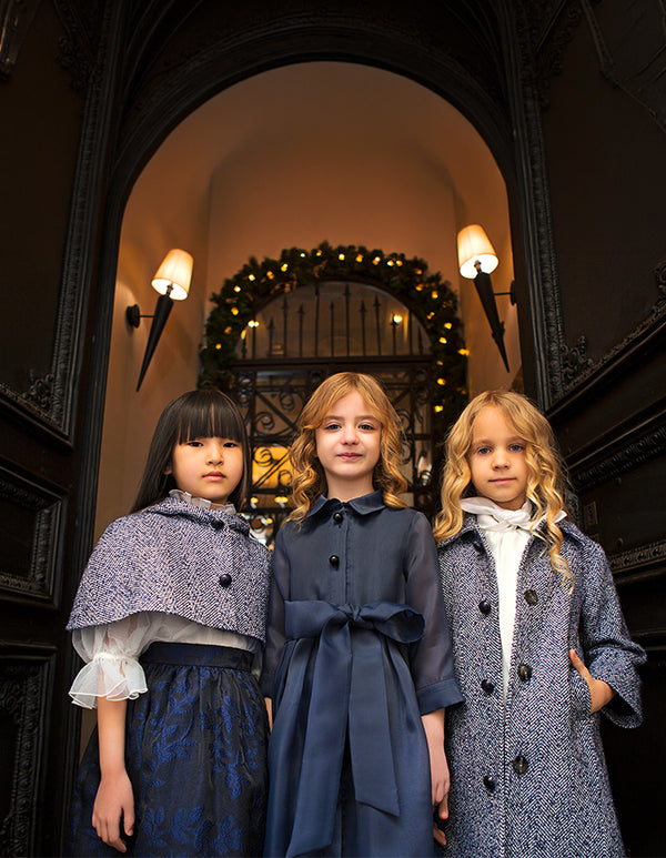 Three girls model navy special occasion dresses and tailored blue tweed outerwear in a festive entryway