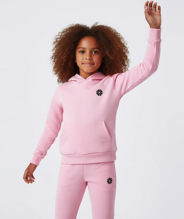 Front-facing lifestyle image of a girl wearing a pink hoodie with matching jogger pants, captured with an arm raised to highlight playful movement, everyday comfort, and active athleisure styling, available in sizes 3–14