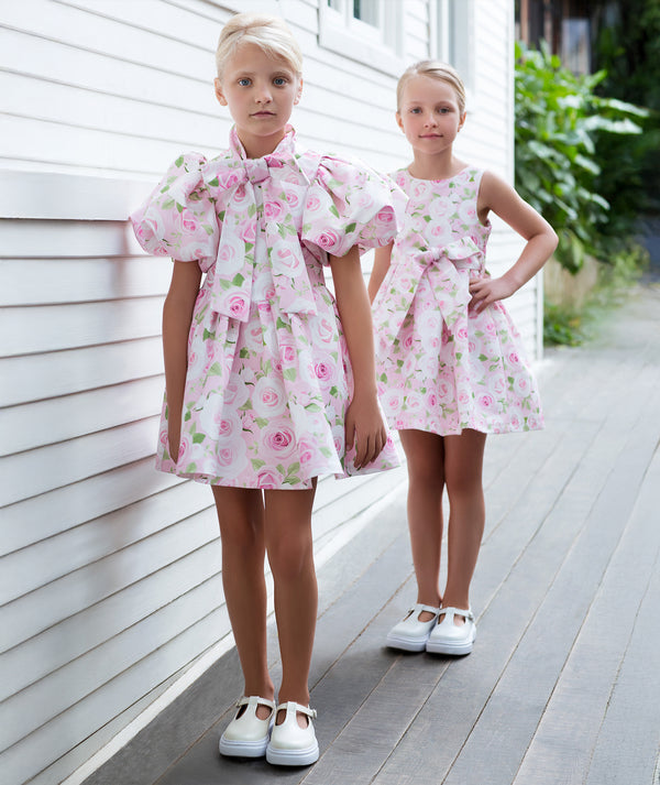Girls pink rose floral dresses with statement bow details and softly structured silhouettes, styled for birthday celebrations, flower girl roles, family gatherings, and elegant summer occasions, available in sizes 3–12