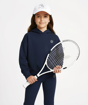 Studio portrait of a girl wearing a navy athletic hoodie and matching leggings from GoldenCubs. She holds a tennis racket and wears a white GoldenCubs cap, representing the brand’s stylish kids’ activewear line inspired by Lululemon.