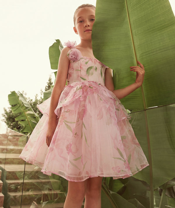 Young girl in a pink floral dress standing among large green leaves.