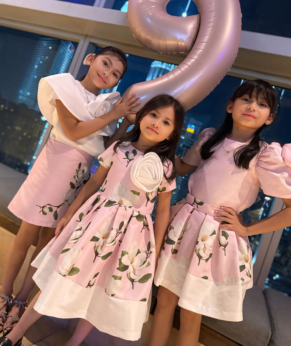 Three young girls in matching dresses with floral patterns, standing indoors with a large number two balloon.