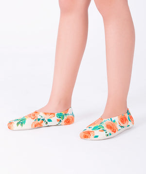Girl wearing orange floral swim shoes by Mama Luma. The slip-on design features bright orange rose prints on a soft white background, ideal for pool, beach, or summer vacations.