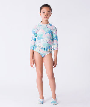Girl wearing a pastel printed long-sleeve rash guard swimsuit by Mama Luma, featuring ruffle details at the waist and matching swim shoes. The design includes soft pink, mint, and blue tones. Perfect for beach days, poolside fun, and summer vacations. Available in girls’ sizes 3–12 years.