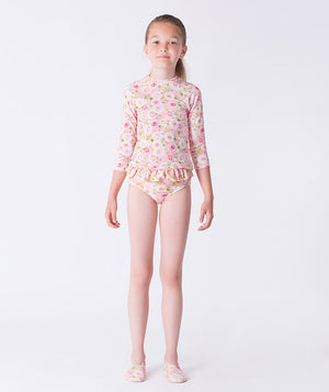 Girl wearing a Mama Luma long-sleeve rash guard swimsuit with pink floral print, featuring ruffle details at the waist and matching floral swim shoes. Perfect for poolside fun, beach days, and summer vacations. Available in girls’ sizes 3–12 years.