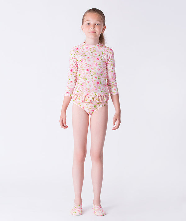 Girl wearing a Mama Luma long-sleeve rash guard swimsuit with pink floral print, featuring ruffle details at the waist and matching floral swim shoes. Perfect for poolside fun, beach days, and summer vacations. Available in girls’ sizes 3–12 years.