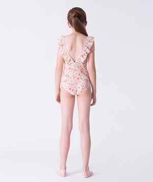 Girl seen from the back wearing a Mama Luma one-piece pink floral swimsuit with ruffle shoulder straps and a low V-back design. Styled with matching floral swim shoes. Perfect for summer vacations, pool days, and beach adventures. Available in girls’ sizes 3–12 years.