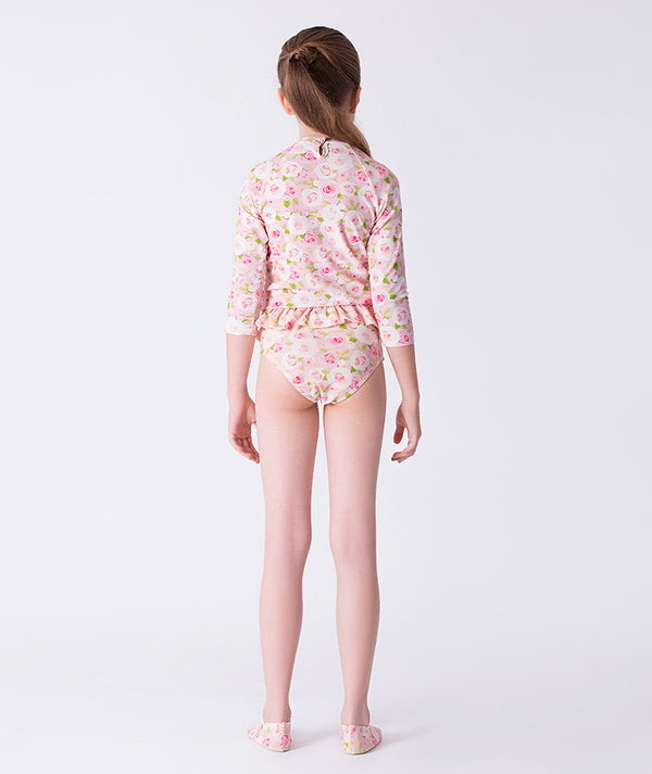 Girl seen from the back wearing a Mama Luma long-sleeve rash guard swimsuit in pink floral print, featuring a ruffled waist and matching swim shoes. Perfect for pool days, beach vacations, and summer fun. Available in girls’ sizes 3–12 years.