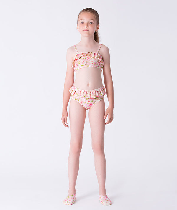 Girl wearing a Mama Luma pink floral two-piece bikini with ruffle trim and matching floral swim shoes. A charming summer swimwear look perfect for poolside, beach days, and vacations. Available in girls’ sizes 3–12 years.