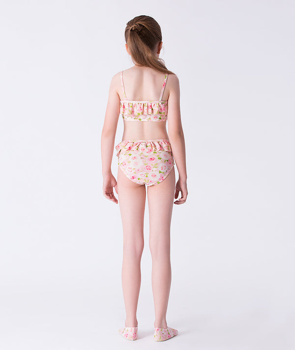 Girl seen from the back wearing a Mama Luma two-piece pink floral bikini with ruffle details and matching floral swim shoes. A charming summer look perfect for the beach, pool, and vacation days. Available in girls’ sizes 3–12 years.