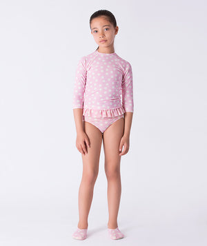 Girl wearing a Mama Luma pink polka-dot long-sleeve rash guard swimsuit with ruffle waist detail and matching swim shoes. A playful and elegant design for summer adventures, pool days, and beach vacations. Available in girls’ sizes 3–12 years.