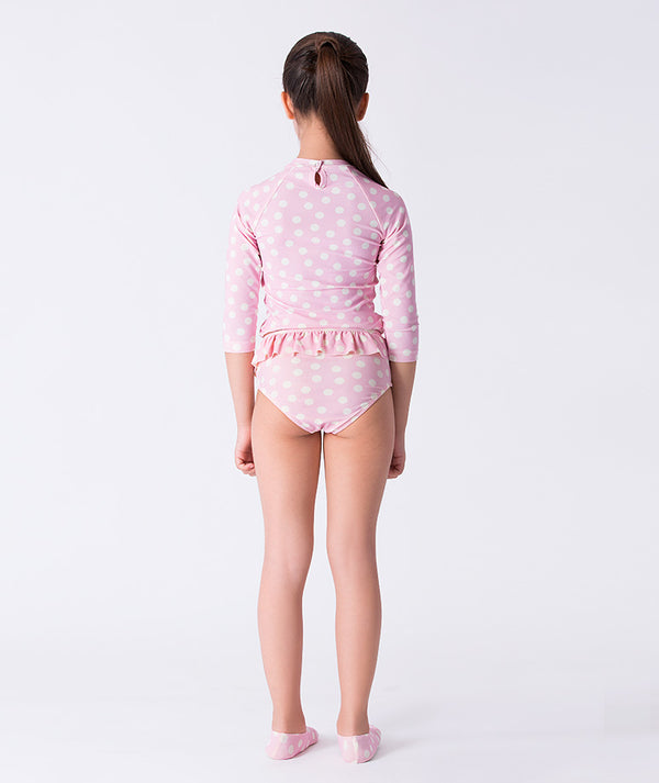 Girl seen from the back wearing a Mama Luma pink polka-dot long-sleeve rash guard swimsuit with ruffle waist and matching swim shoes. Perfect for pool days, beach vacations, and summer fun. Available in girls’ sizes 3–12 years.