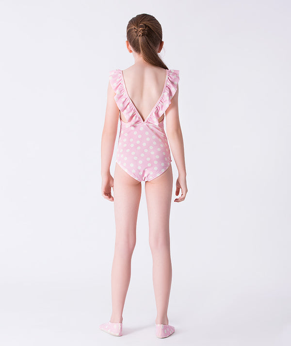 Girl seen from the back wearing a Mama Luma pink polka-dot one-piece swimsuit with ruffle shoulder straps and low V-back design, styled with matching polka-dot swim shoes. Perfect for pool days, beach vacations, and summer holidays. Available in girls’ sizes 3–12 years.