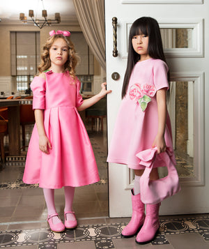 Two girls wearing matching bright pink dresses with floral appliqués, pink boots and coordinated handbags