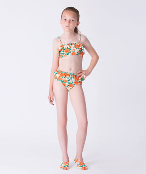 Girl wearing a Mama Luma orange floral two-piece bikini with ruffle trim and matching floral swim shoes. A bright, cheerful swimsuit ideal for summer vacations, pool days, and beach outings. Available in girls’ sizes 3–12 years.
