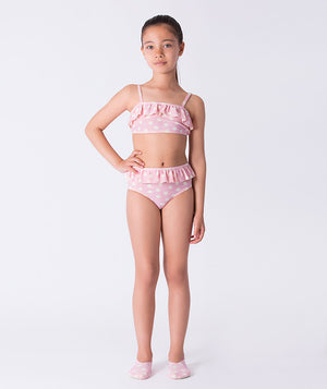 Girl wearing a Mama Luma pink polka-dot two-piece bikini with ruffle trim and matching swim shoes. A playful pastel swimsuit ideal for summer vacations, pool days, and beach fun. Available in girls’ sizes 3–12 years.