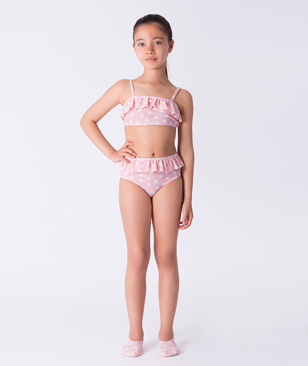 Girl wearing a Mama Luma pink polka-dot two-piece bikini with ruffle trim and matching swim shoes. A playful pastel swimsuit ideal for summer vacations, pool days, and beach fun. Available in girls’ sizes 3–12 years.