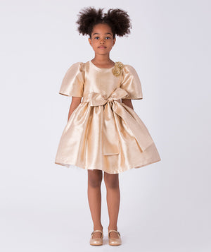 Front view of gold metallic girls dress with puff sleeves, floral shoulder embellishment and statement waist bow.