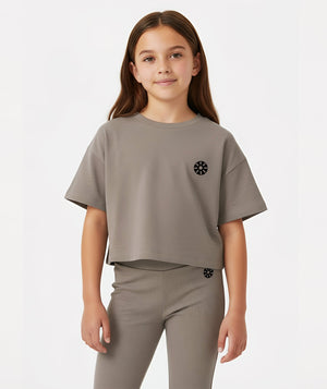 Front-facing lifestyle image of a girl wearing a taupe cropped short sleeve t-shirt layered over matching ribbed pants, styled for contemporary everyday outfits, school comfort, and relaxed movement, available in sizes 3–14