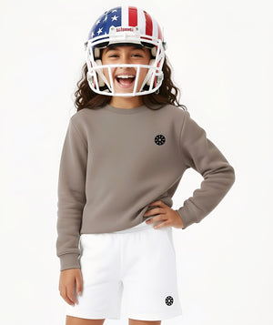 Front-facing lifestyle image of a girl wearing a taupe long sleeve sweatshirt and white shorts with a football helmet, styled for playful sports energy, bold athleisure looks, and confident everyday wear, available in sizes 3–14