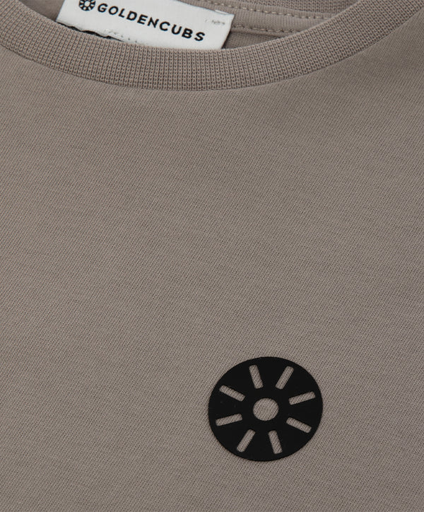 Close-up detail of a taupe girls short sleeve t-shirt featuring a minimalist black chest emblem and soft knit texture, designed for everyday wear, school outfits, and casual looks, available in sizes 3–14