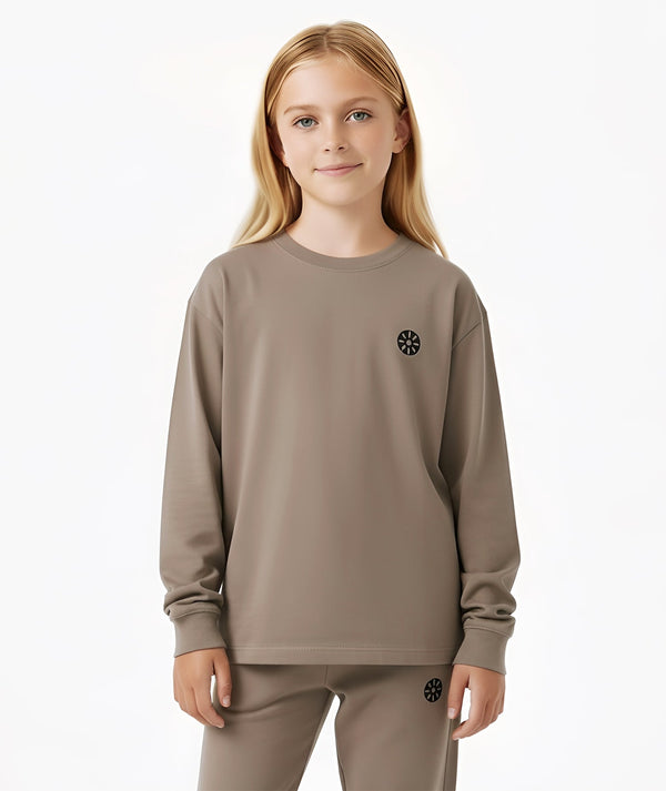 Front-facing lifestyle image of a girl wearing a taupe long sleeve top with a relaxed fit and subtle chest emblem, styled for calm everyday outfits, school layering, and comfortable daily wear, available in sizes 3–14