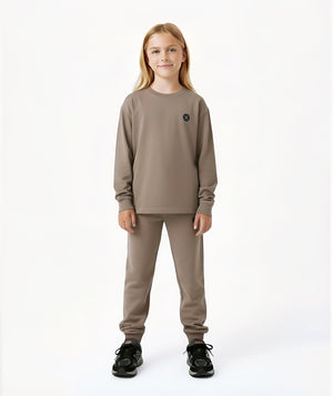 Front-facing lifestyle image of a girl wearing a taupe long sleeve sweatshirt with coordinating pants, styled for clean everyday outfits, school-ready comfort, and relaxed daily movement, available in sizes 3–14