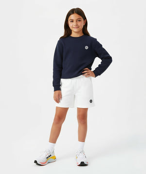 Front-facing lifestyle image of a girl wearing a navy long sleeve sweatshirt with white shorts, styled for transitional weather outfits, casual school days, and relaxed movement, available in sizes 3–14