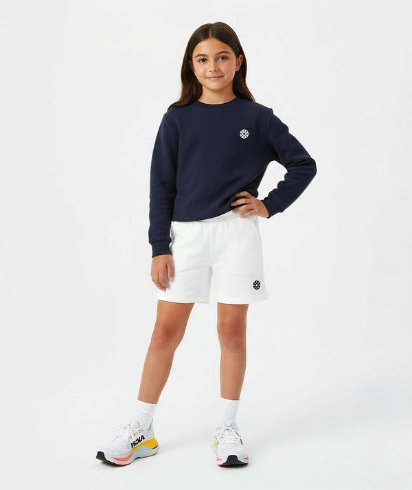 Front-facing lifestyle image of a girl wearing a navy long sleeve sweatshirt with white shorts, styled for transitional weather outfits, casual school days, and relaxed movement, available in sizes 3–14