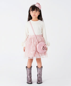 A young girl wearing a white long-sleeve blouse featuring rosette flower details on the cuffs, paired with a pink floral tulle skirt and western boots.