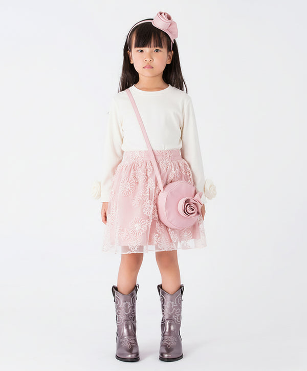 A young girl wearing a white long-sleeve blouse featuring rosette flower details on the cuffs, paired with a pink floral tulle skirt and western boots.